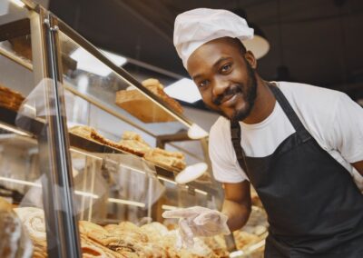 Cheerful baker in traditional attire presents a variety of fresh pastries at a bakery.